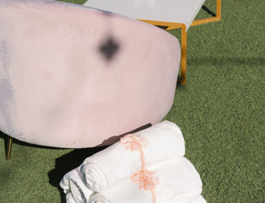 Three rolled white towels tied with pink ribbon are stacked on green grass next to a white table with two glasses and a curved, light pink chair, creating a serene setting perfect for an elegant wedding or spring wedding at Cielo Farms.