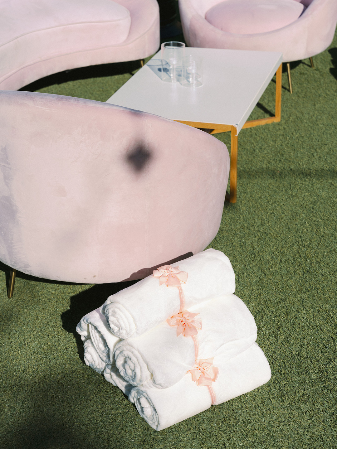 Three rolled white towels tied with pink ribbon are stacked on green grass next to a white table with two glasses and a curved, light pink chair, creating a serene setting perfect for an elegant wedding or spring wedding at Cielo Farms.