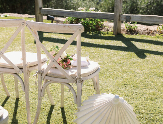 Two white wooden chairs with small flower bouquets on the seats sit on grass at Cielo Farms, next to a white paper parasol and wooden fence, with greenery and hills in the background—a perfect touch for an elegant spring wedding.