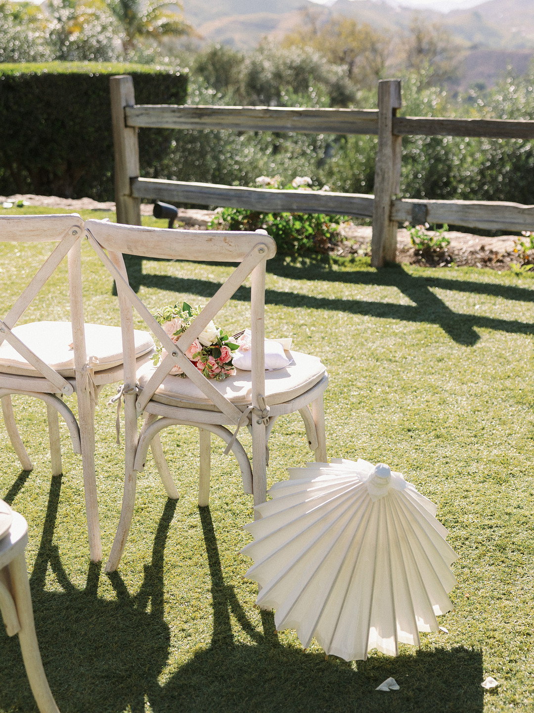 Two white wooden chairs with small flower bouquets on the seats sit on grass at Cielo Farms, next to a white paper parasol and wooden fence, with greenery and hills in the background—a perfect touch for an elegant spring wedding.