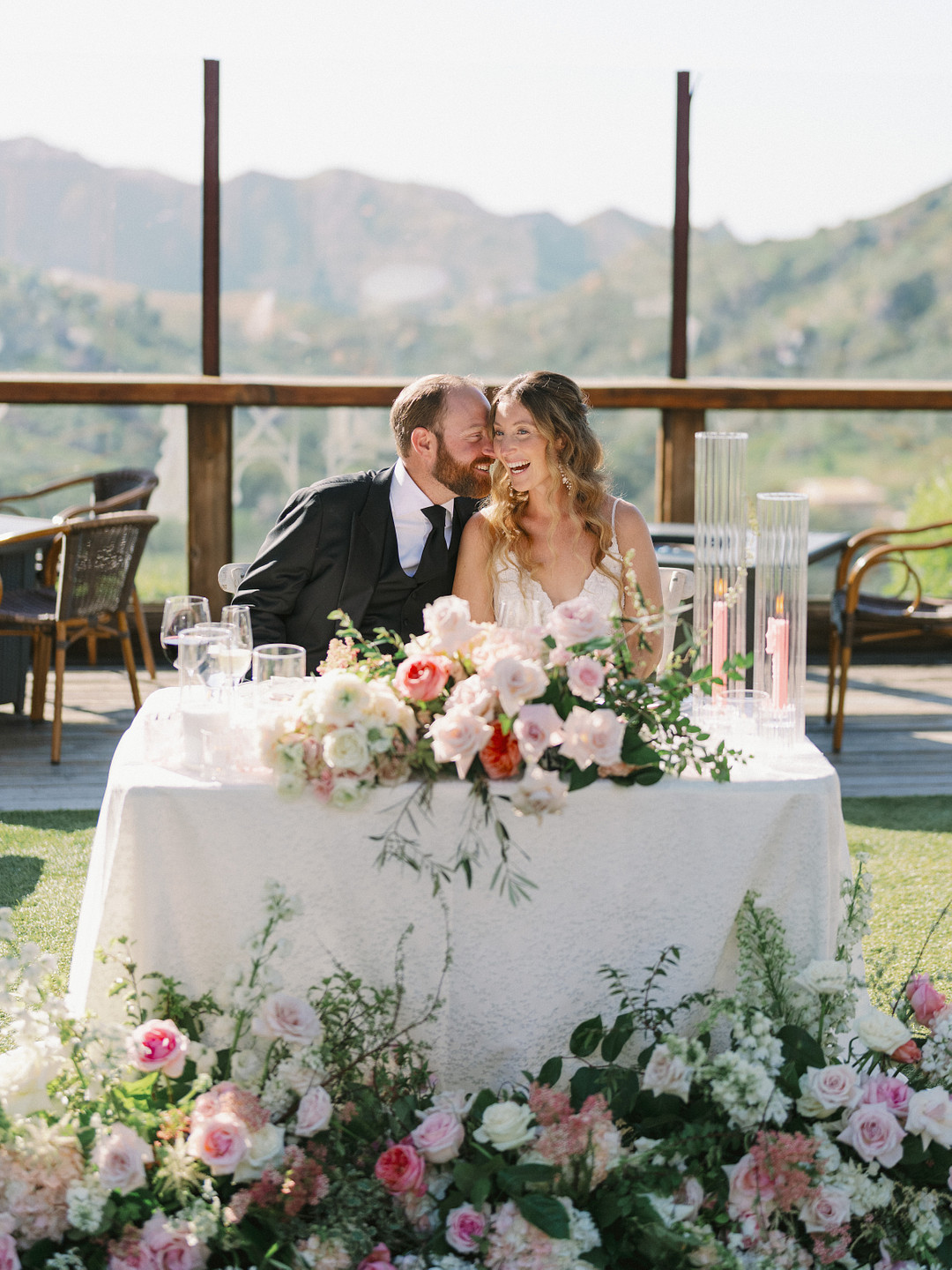 A newlywed couple sits at a flower-adorned table outdoors at Cielo Farms, with the groom leaning in to whisper to the bride. Elegant wedding details surround them, with mountains and clear skies in the background.