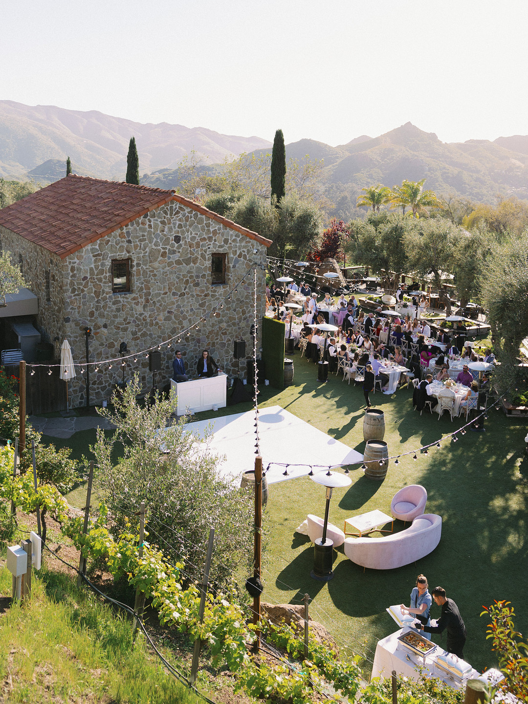Elegant wedding at Cielo Farms, with guests seated at tables by a stone building, surrounded by lush greenery and mountains under clear spring skies.