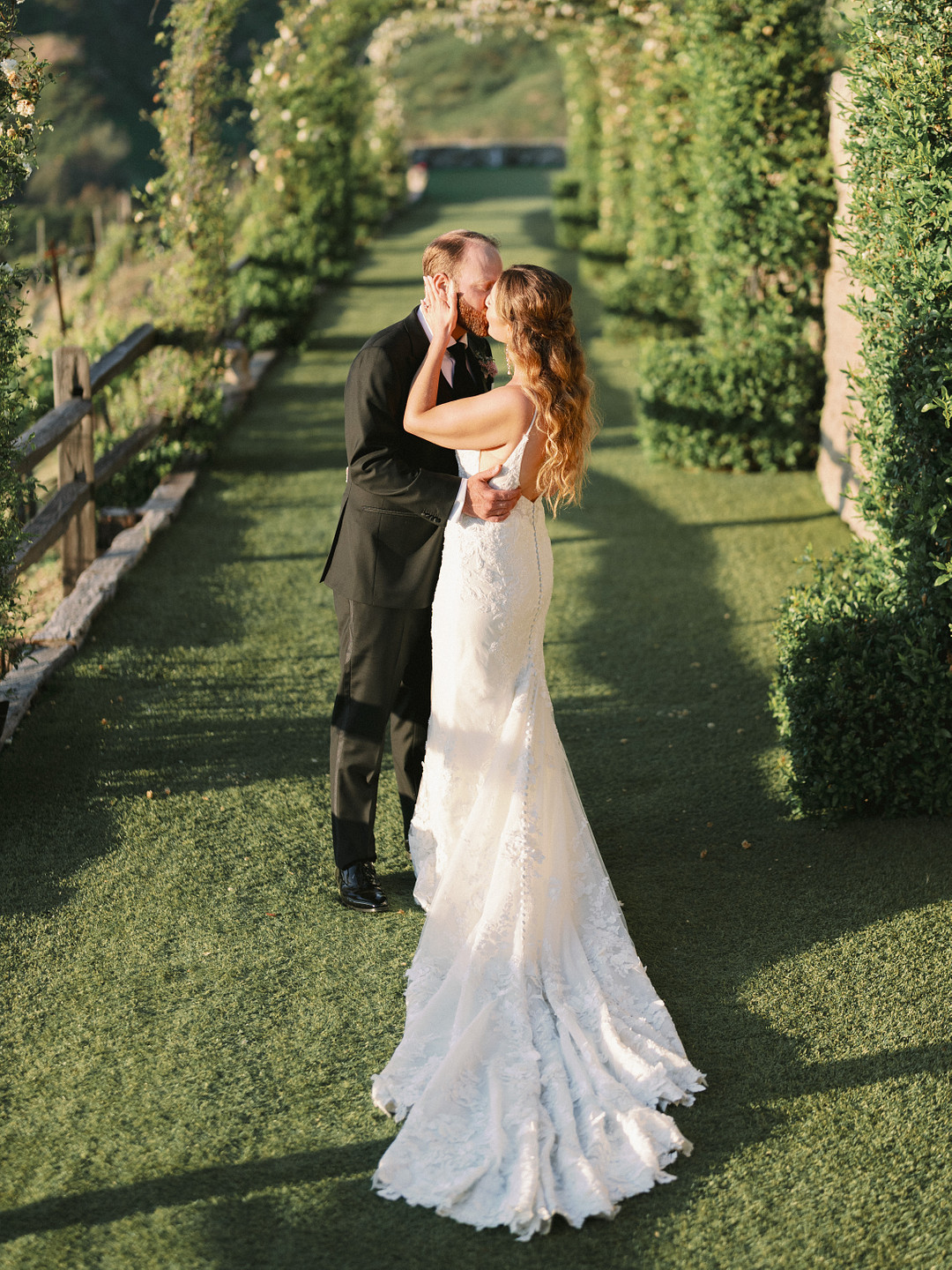 An elegant wedding at Cielo Farms: a bride and groom kiss outdoors on a grassy aisle lined with greenery and arches, sunlight casting long shadows.