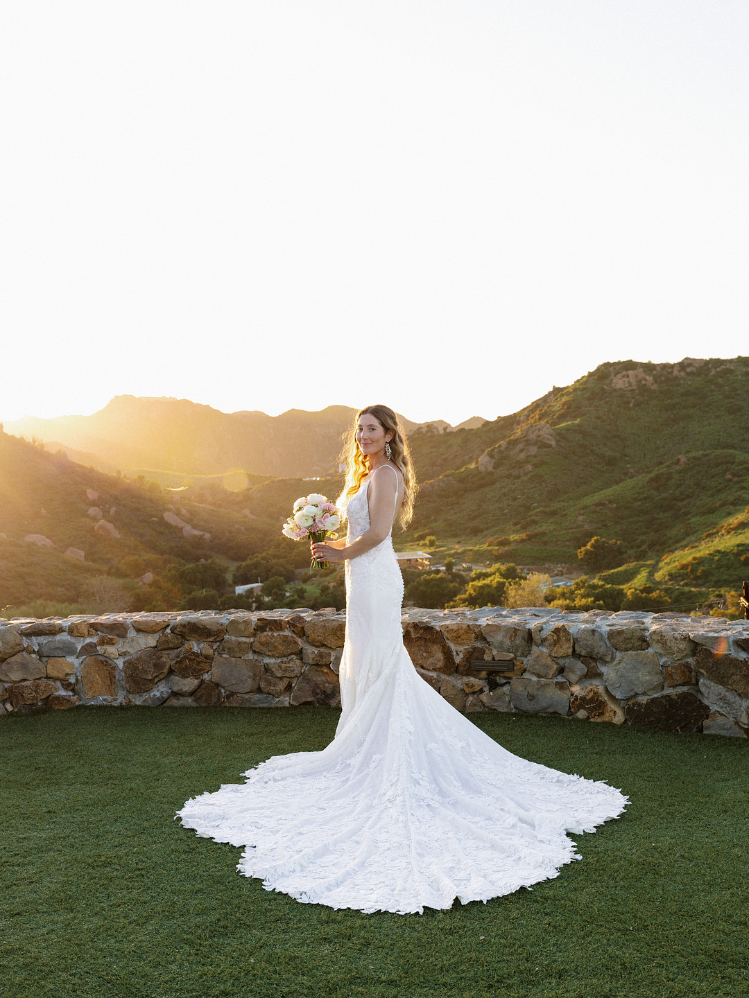 A bride in a white lace wedding dress holding a bouquet stands outdoors on grass at Cielo Farms, with mountains and sunlight in the background—capturing the essence of an elegant wedding.