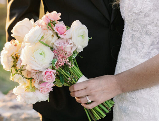 A bride in a white lace dress holds a bouquet of white and pink flowers, standing next to a person in a black suit at an elegant wedding, capturing the charm of a spring wedding at Cielo Farms.