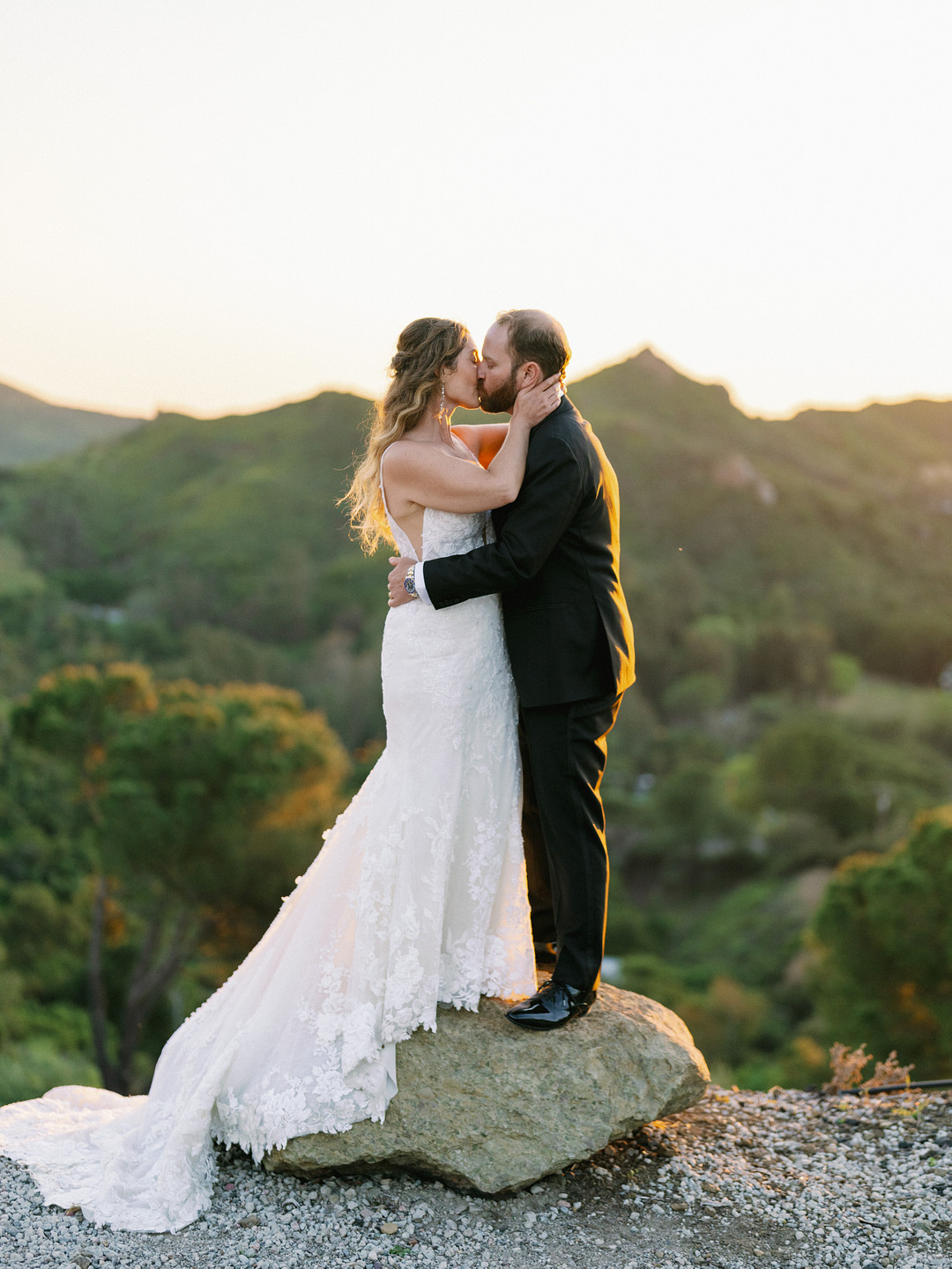 A bride and groom in elegant wedding attire stand on a rock, embracing and kissing, with green hills and trees in the background at sunset for their romantic Spring Wedding.
