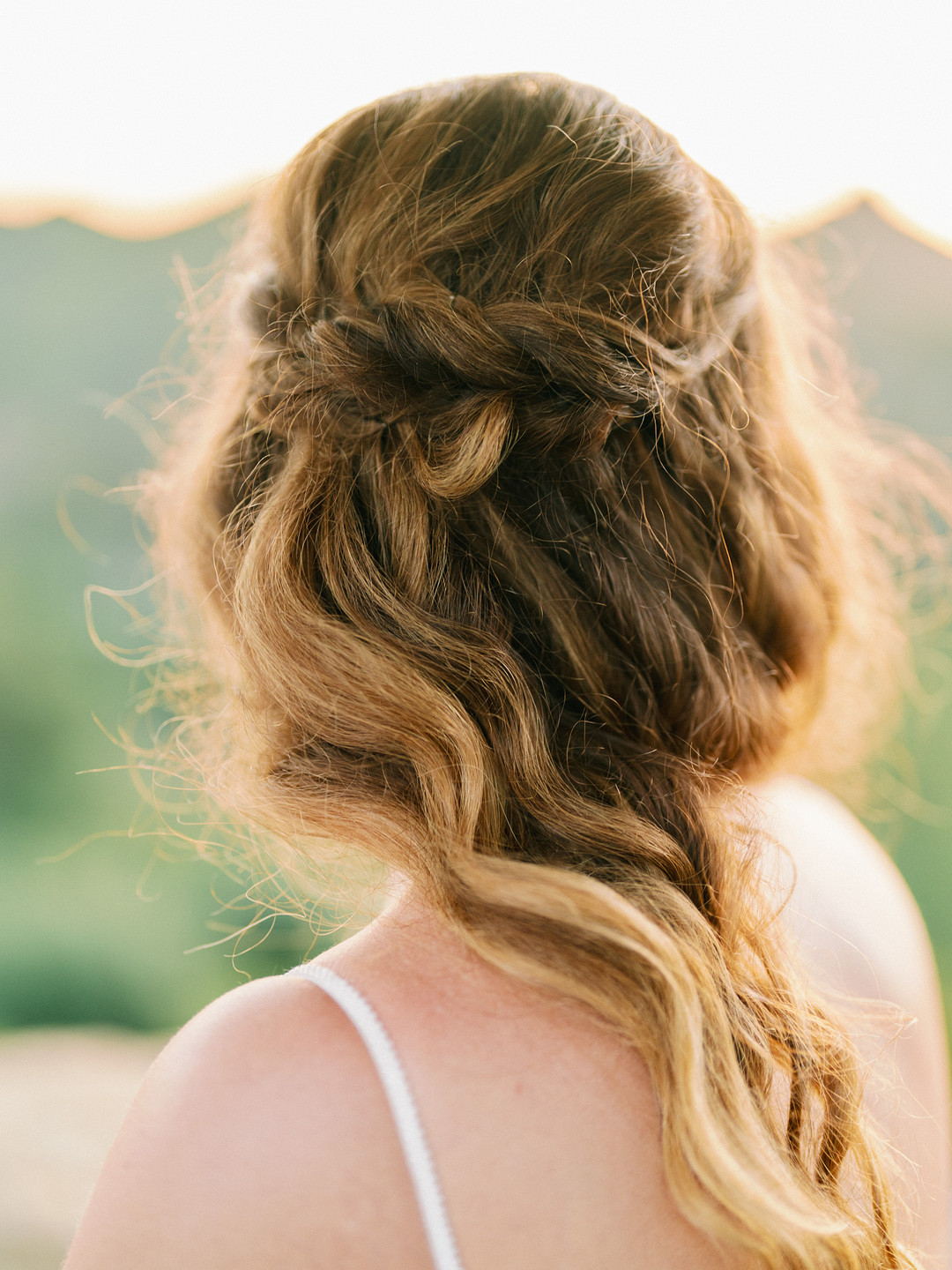 Person with long, wavy, light brown hair styled in loose braids, photographed from behind outdoors in soft natural light at a picturesque Cielo Farms spring wedding.