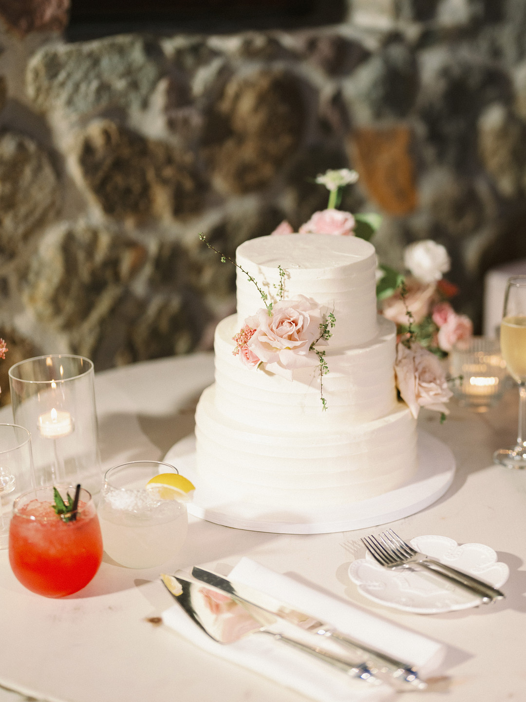 A two-tier white frosted cake with pink flowers sits on a table next to two cocktails, a lit candle, and place settings, creating an elegant wedding vibe against a stone wall backdrop at Cielo Farms.