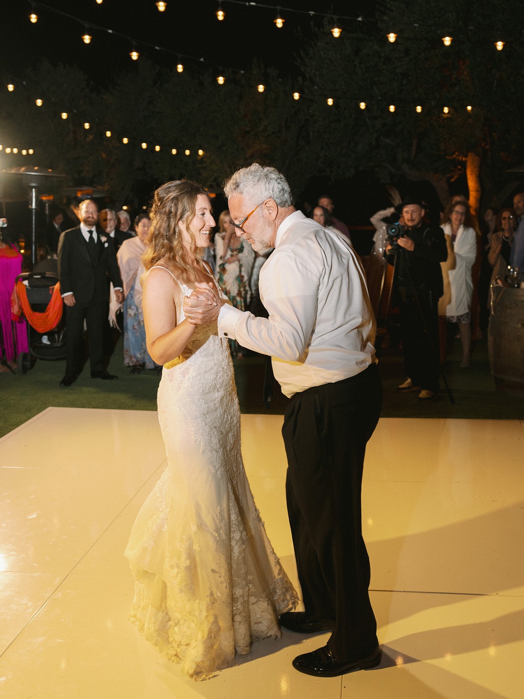 A bride in a white dress dances with an older man in a white shirt and black pants on an outdoor dance floor at Cielo Farms, surrounded by guests celebrating an elegant spring wedding under the night sky.