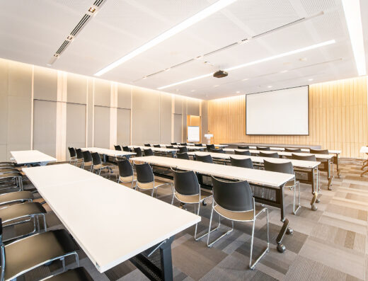 A bright, modern conference room with rows of tables and chairs facing a projector screen and podium.