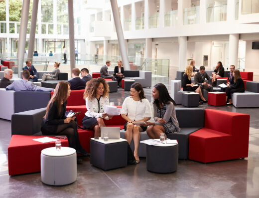 People in business attire sitting on modern couches in an open, bright office lobby; some are talking in groups, others are using laptops or tablets.