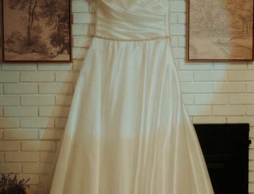 A white satin wedding dress on a hanger is displayed against a white brick wall between two framed landscape drawings.