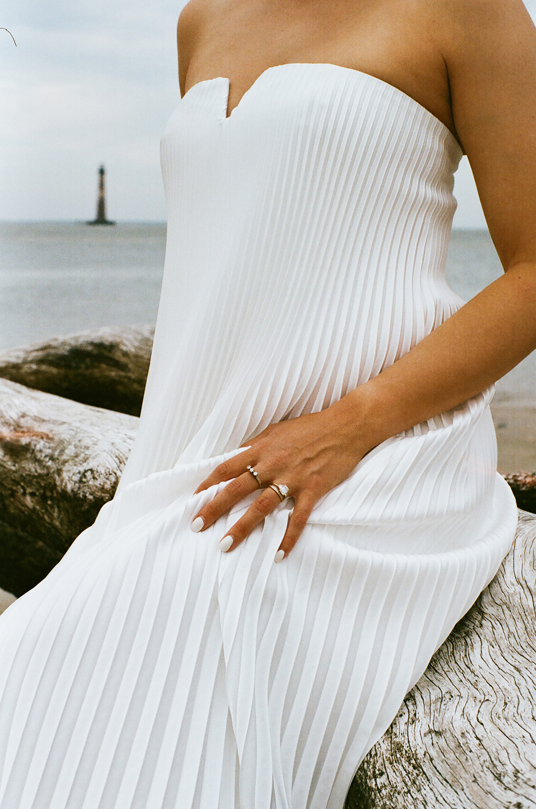Person in a strapless, white, pleated dress sits on a driftwood log by the water, wearing rings and with manicured nails. A distant lighthouse is visible on the horizon.