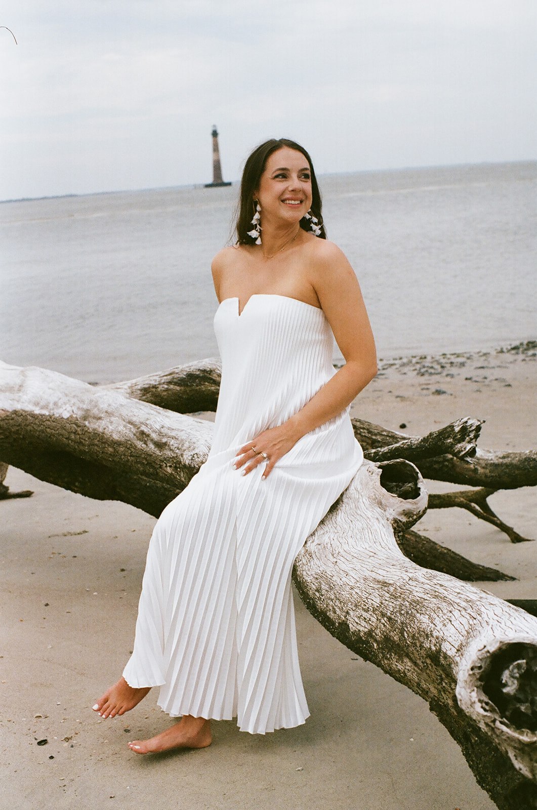 Woman in a strapless white dress sits barefoot on a large driftwood log on a sandy beach, with a lighthouse visible in the background.