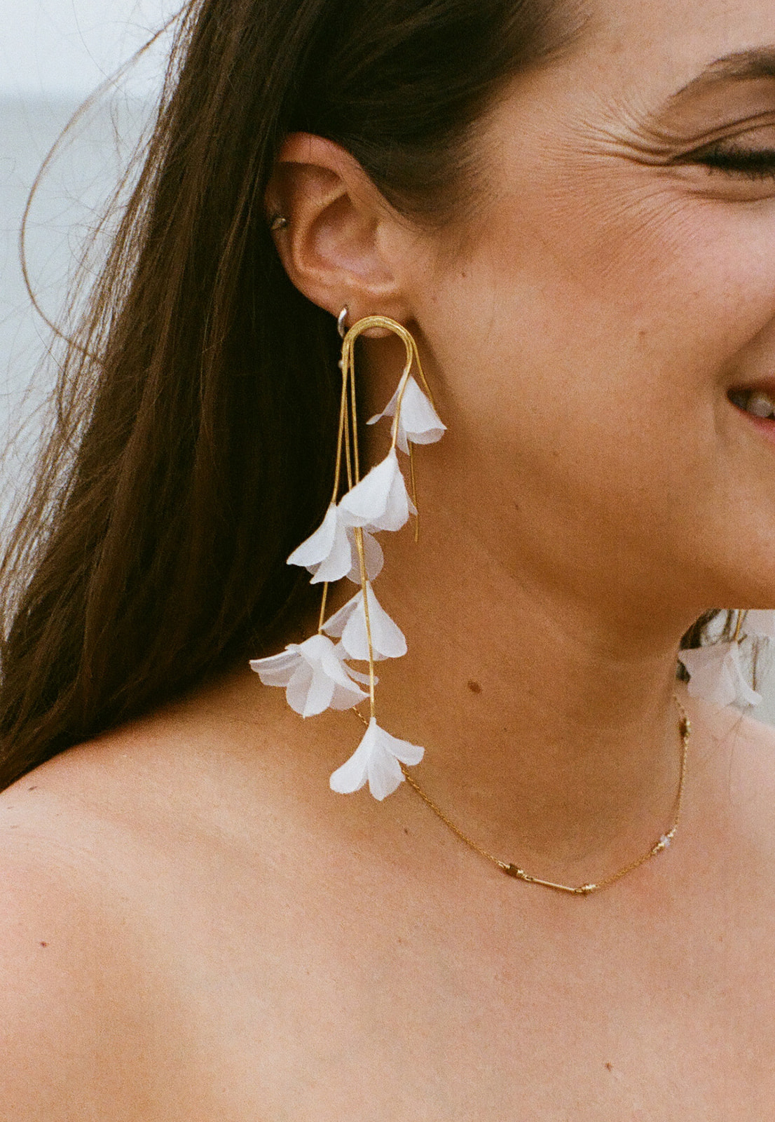 A woman wearing a large gold earring with cascading white flower petals and a delicate gold necklace, smiling, with long brown hair and bare shoulders.