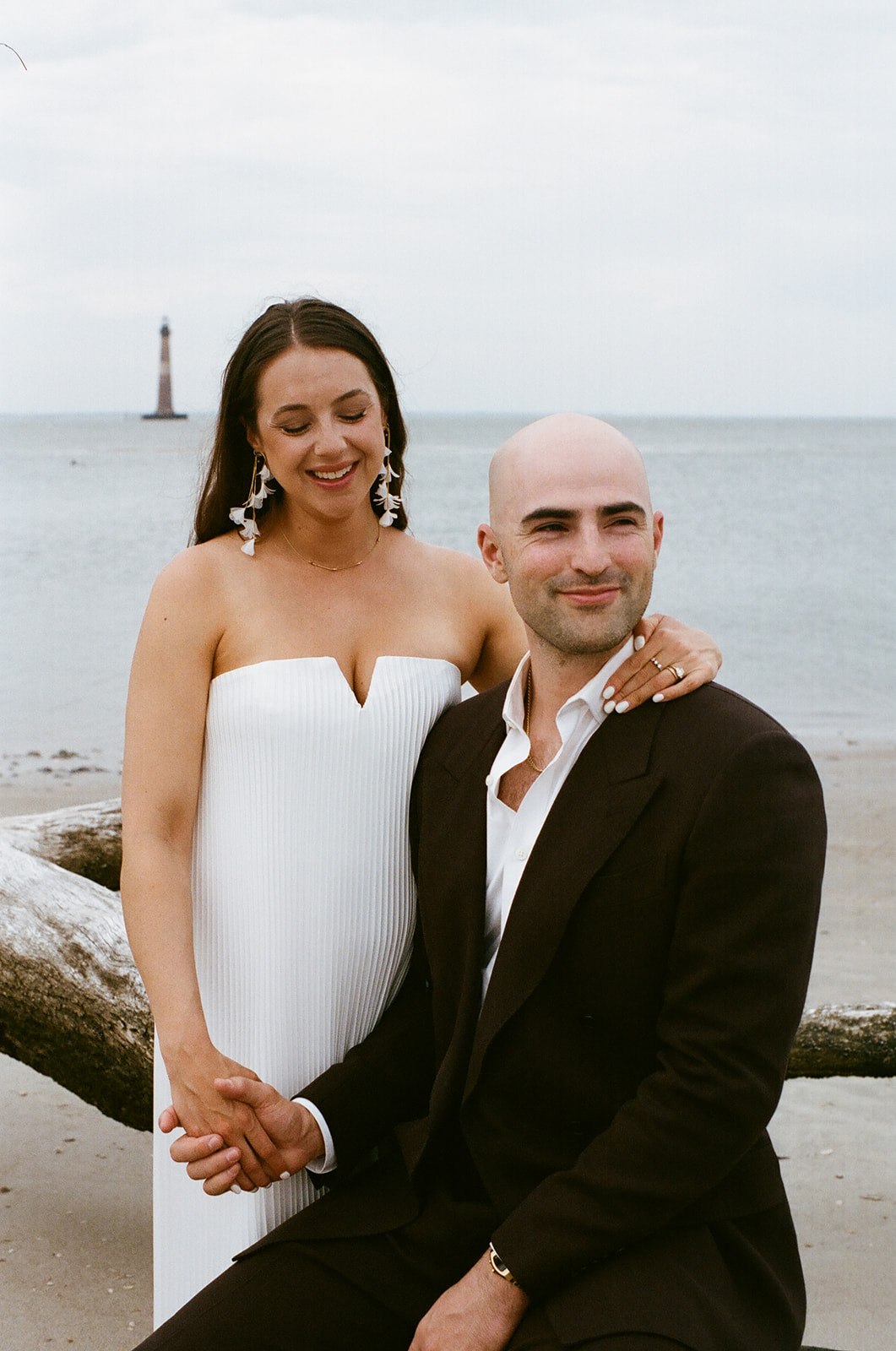A woman in a white dress stands beside a seated man in a black suit on a beach, holding hands, with a lighthouse visible in the background.