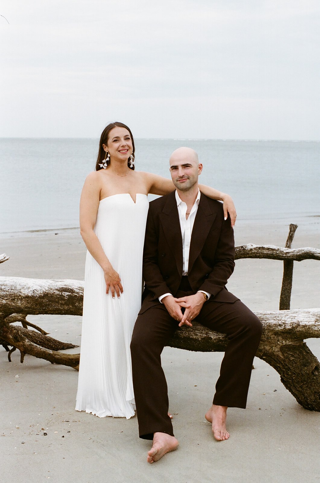 A woman in a white dress stands next to a seated man in a dark suit. They are barefoot on a beach with driftwood, facing the camera, with the ocean in the background.