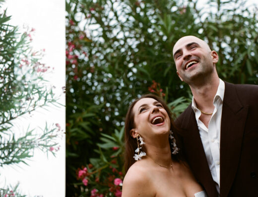 A woman and a man stand close together outdoors, both looking up and smiling, with green foliage and pink flowers in the background.