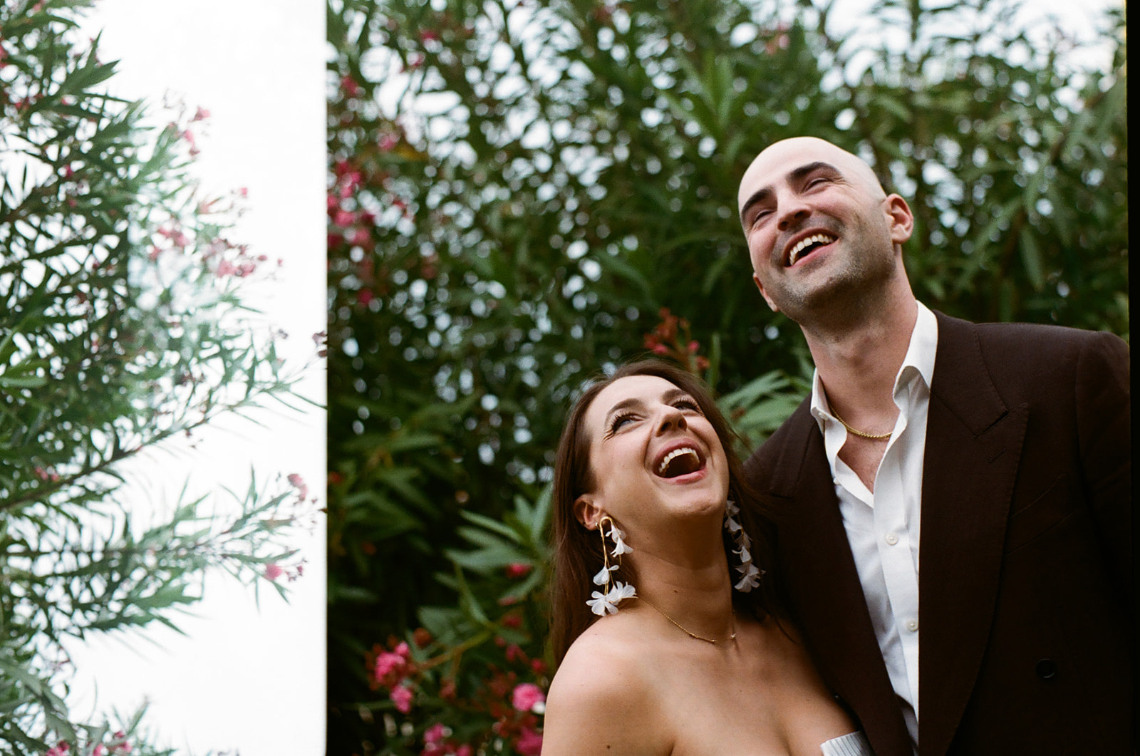 A woman and a man stand close together outdoors, both looking up and smiling, with green foliage and pink flowers in the background.