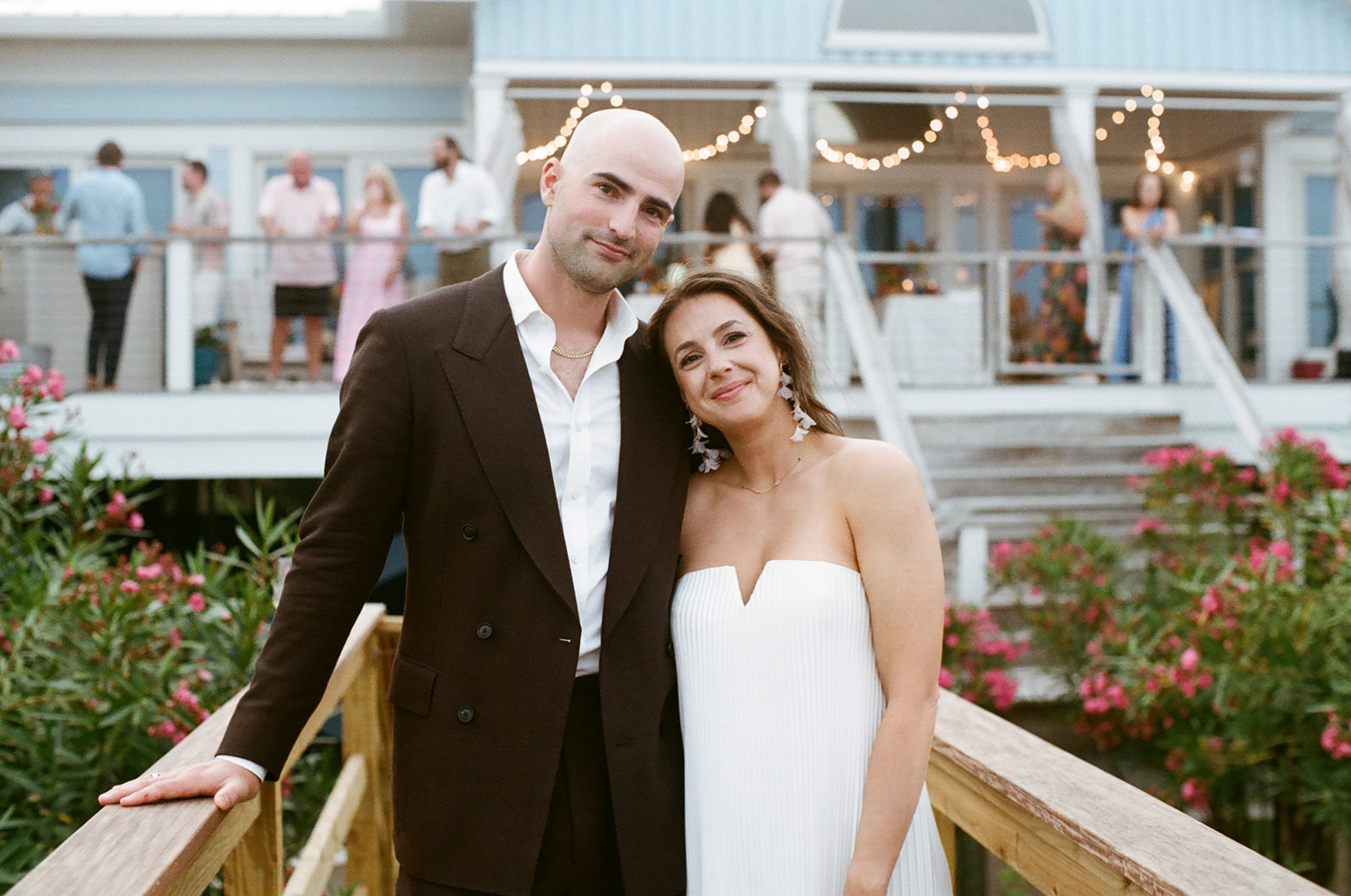 A man in a dark suit and a woman in a white strapless dress pose together on a wooden bridge, with a party and string lights in the background.