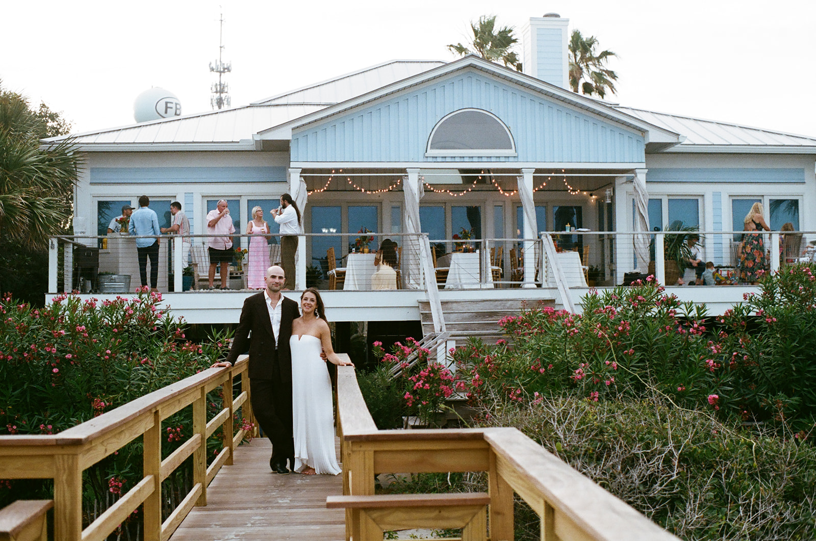 A bride and groom stand on a wooden walkway leading to a blue beach house, with guests mingling on the deck above.