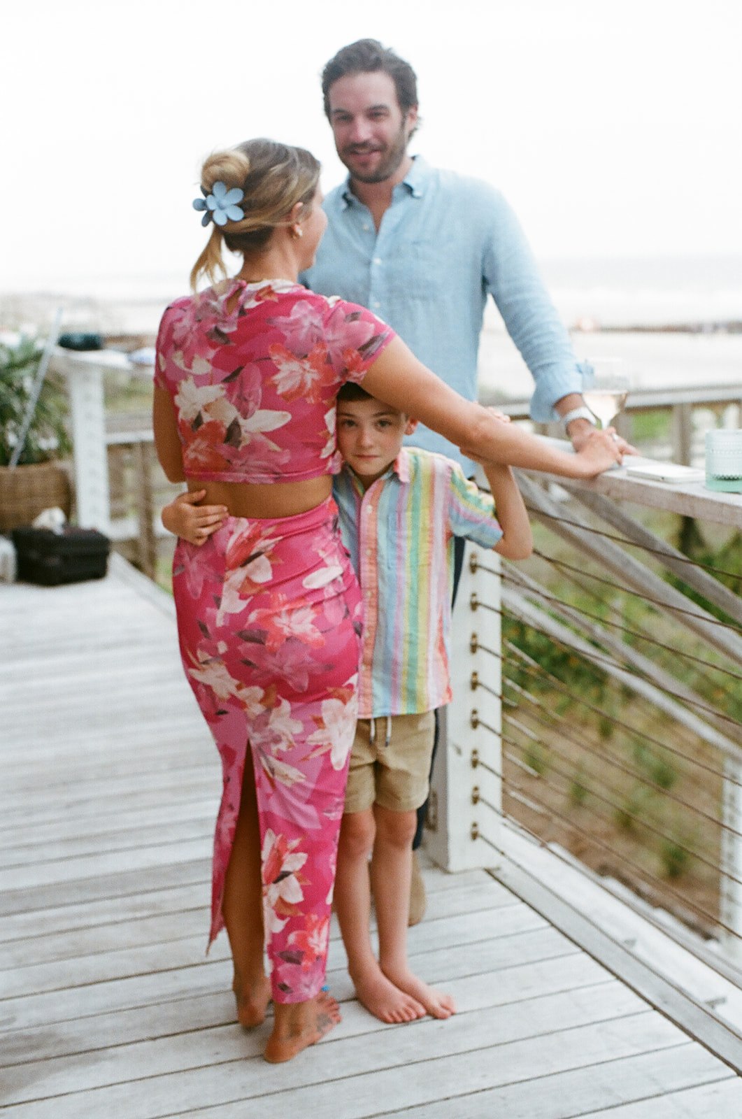 A woman in a pink floral dress hugs a child in a striped shirt and khaki shorts, while a man in a blue shirt stands behind them on a wooden deck.