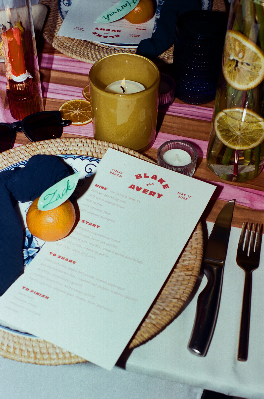 A place setting with a menu, candle, cutlery, napkin, sunglasses, and an orange labeled “Zack” on a woven charger; drinks with lemon slices are in the background.