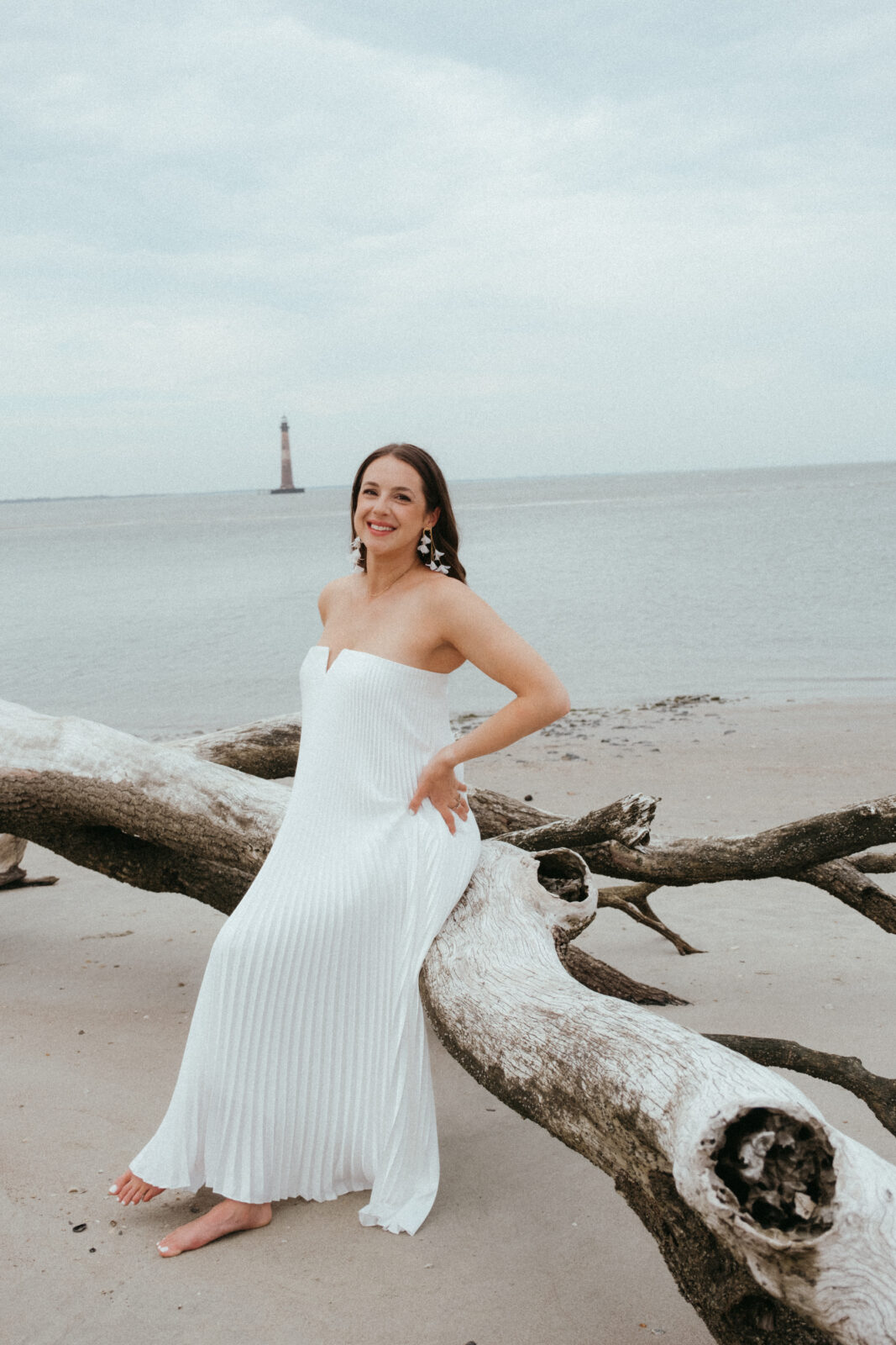 Woman in a white strapless dress sits on a large driftwood log on a sandy beach, with the ocean and a distant lighthouse in the background.