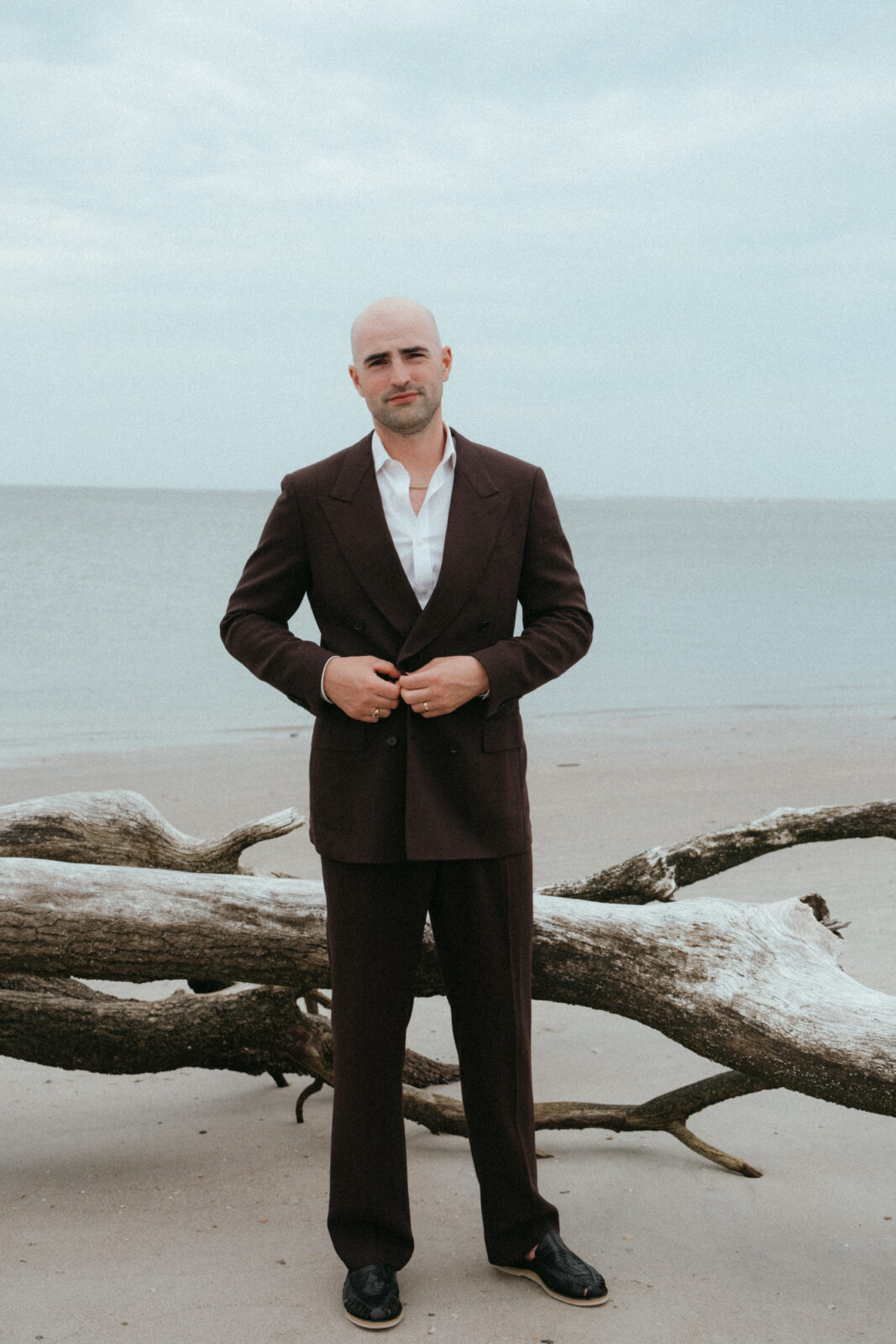 A man in a dark suit stands on a beach in front of a large driftwood log, with the ocean and cloudy sky in the background.