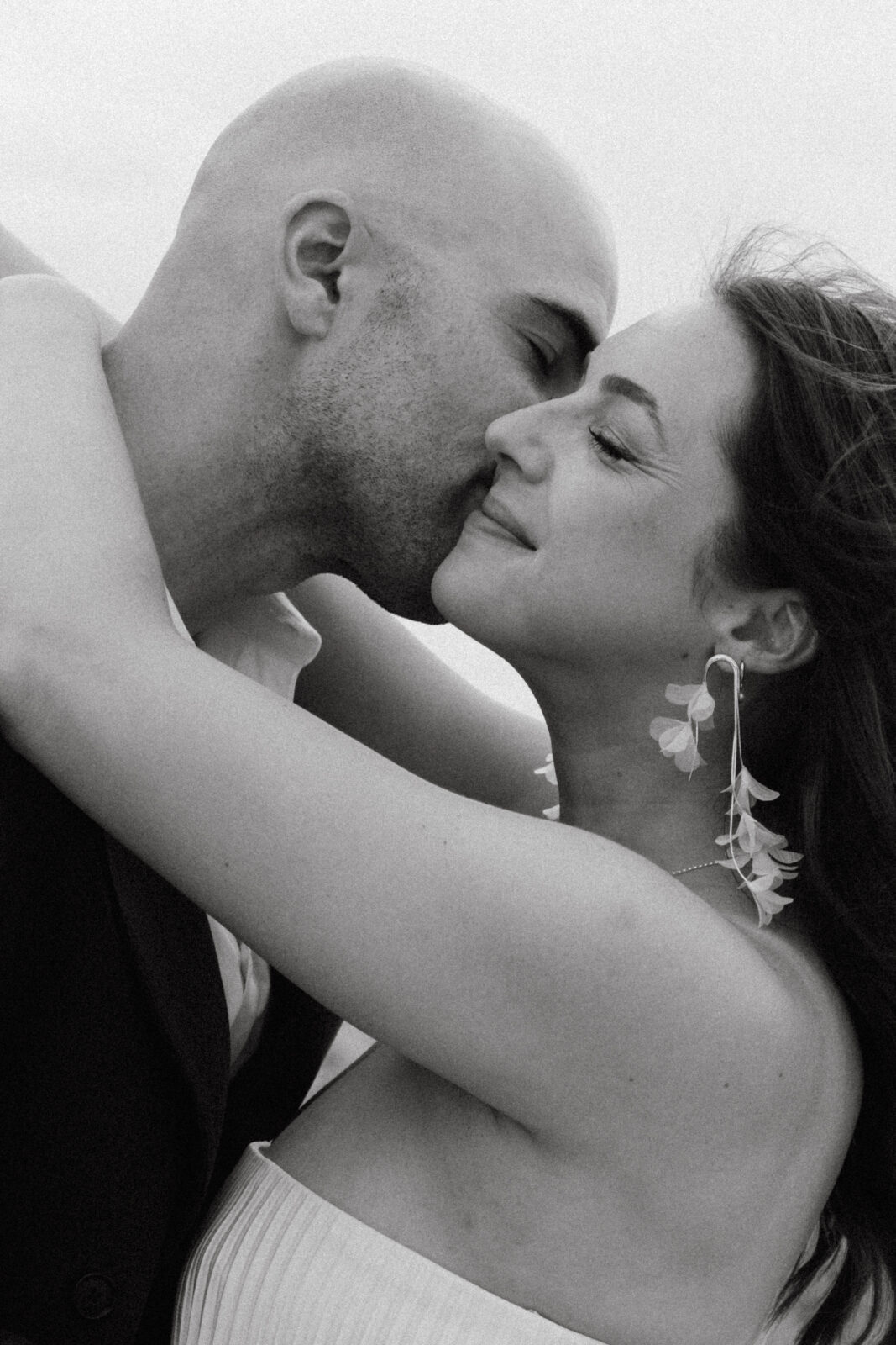 A man gently kisses a woman’s cheek as she smiles with closed eyes, both embracing closely. The image is in black and white.