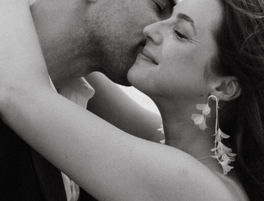 A man gently kisses a woman’s cheek as she smiles with closed eyes, both embracing closely. The image is in black and white.
