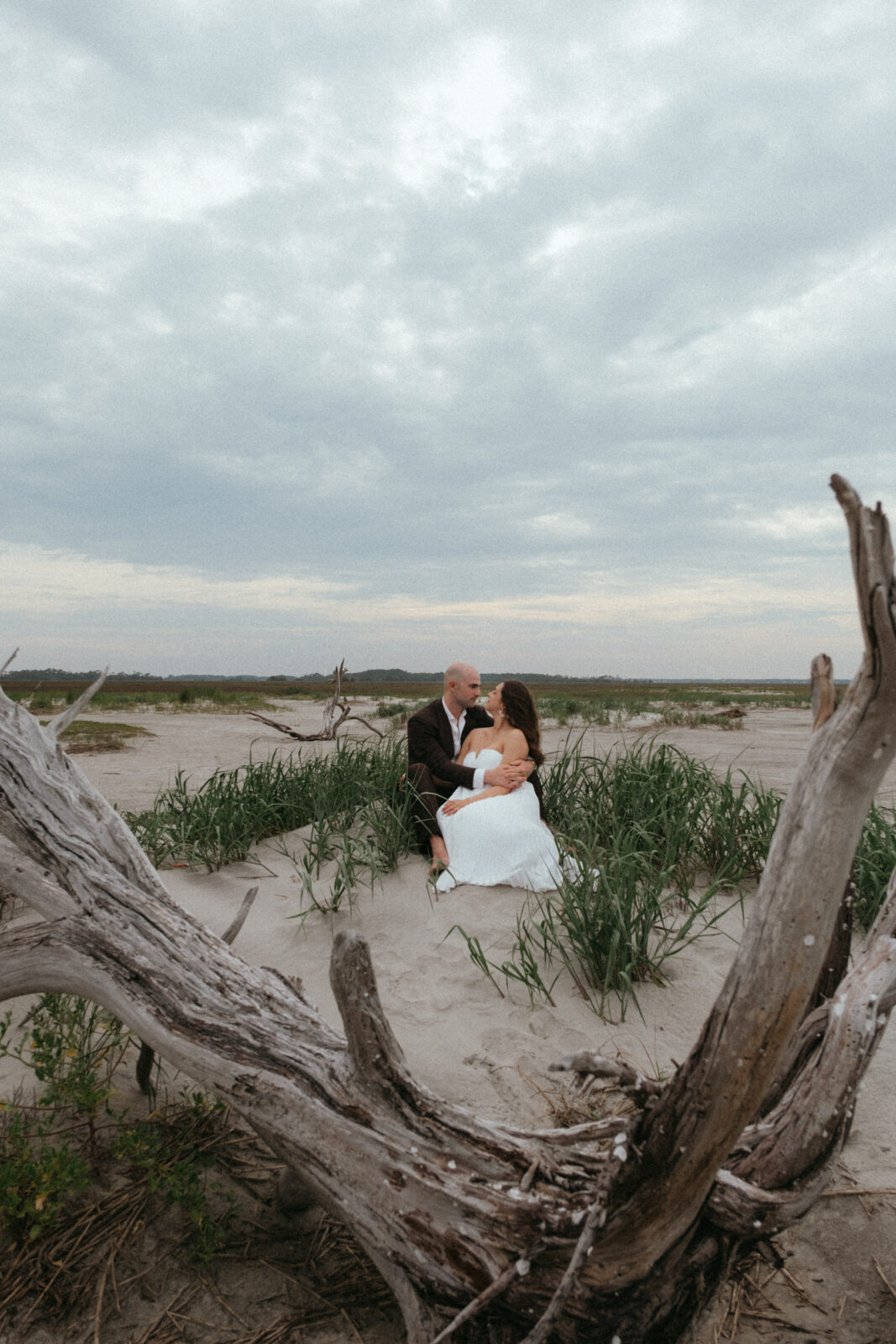 A couple in wedding attire sit close together on a sandy beach, surrounded by driftwood and grass under a cloudy sky.