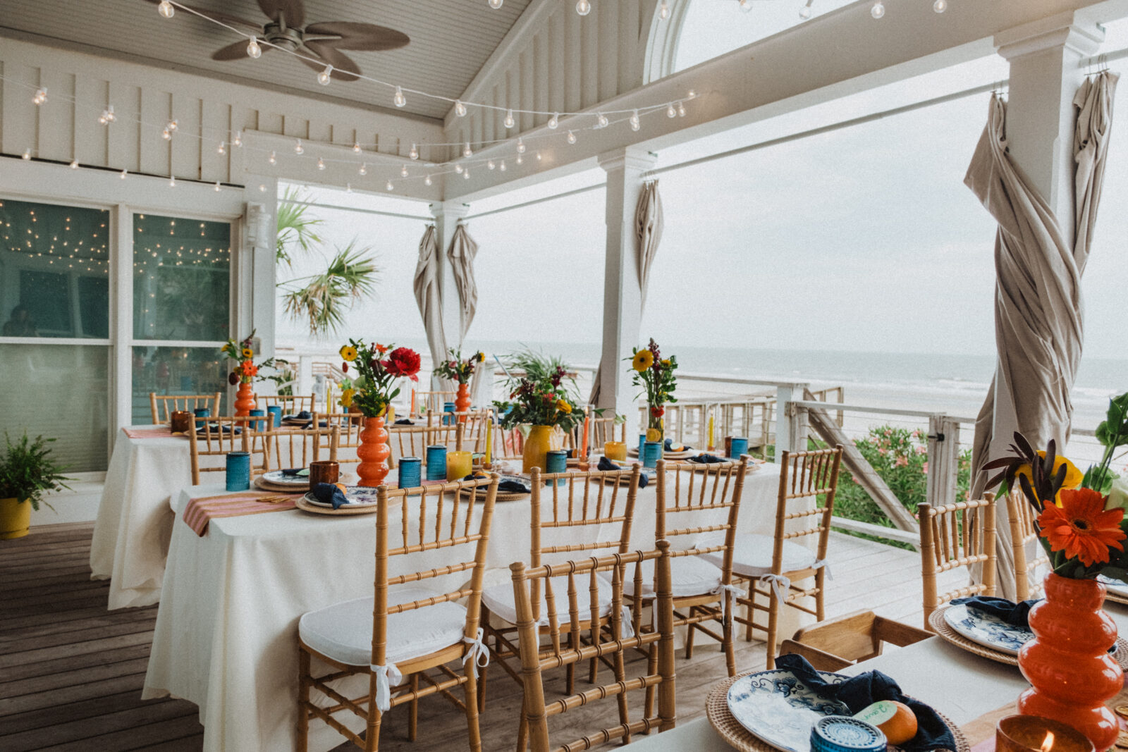 Tables set for an outdoor event on a covered patio overlooking the beach, decorated with flowers, string lights, and wooden chairs.