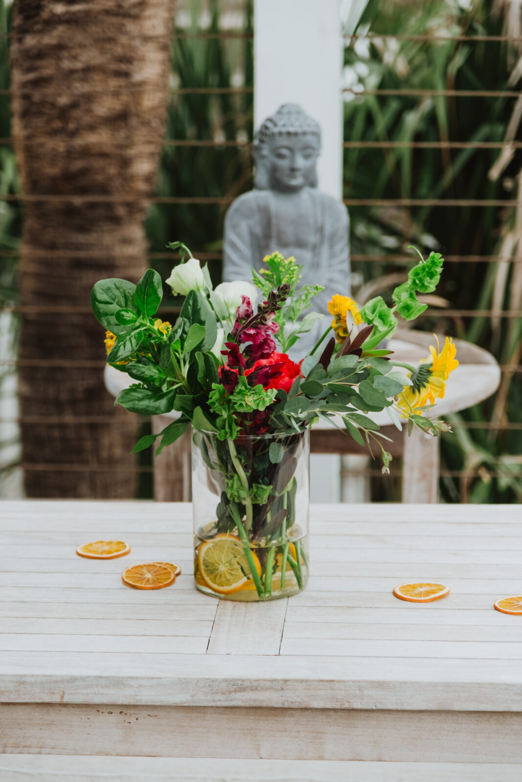 A glass vase with assorted fresh flowers and lemon slices sits on a white wooden table, with dried orange slices nearby. A Buddha statue and greenery are visible in the background.