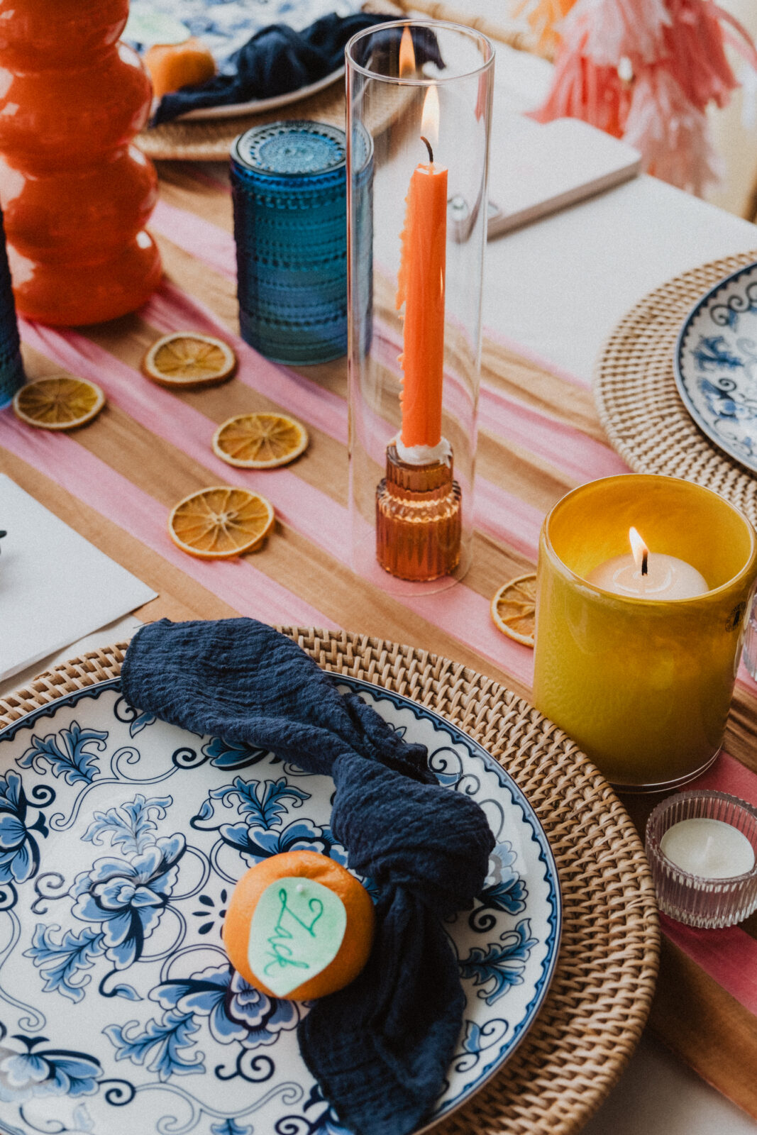 A table setting with blue-patterned plates, a navy napkin, a yellow candle, orange candle in a glass holder, blue glass, and dried orange slices on a striped runner.