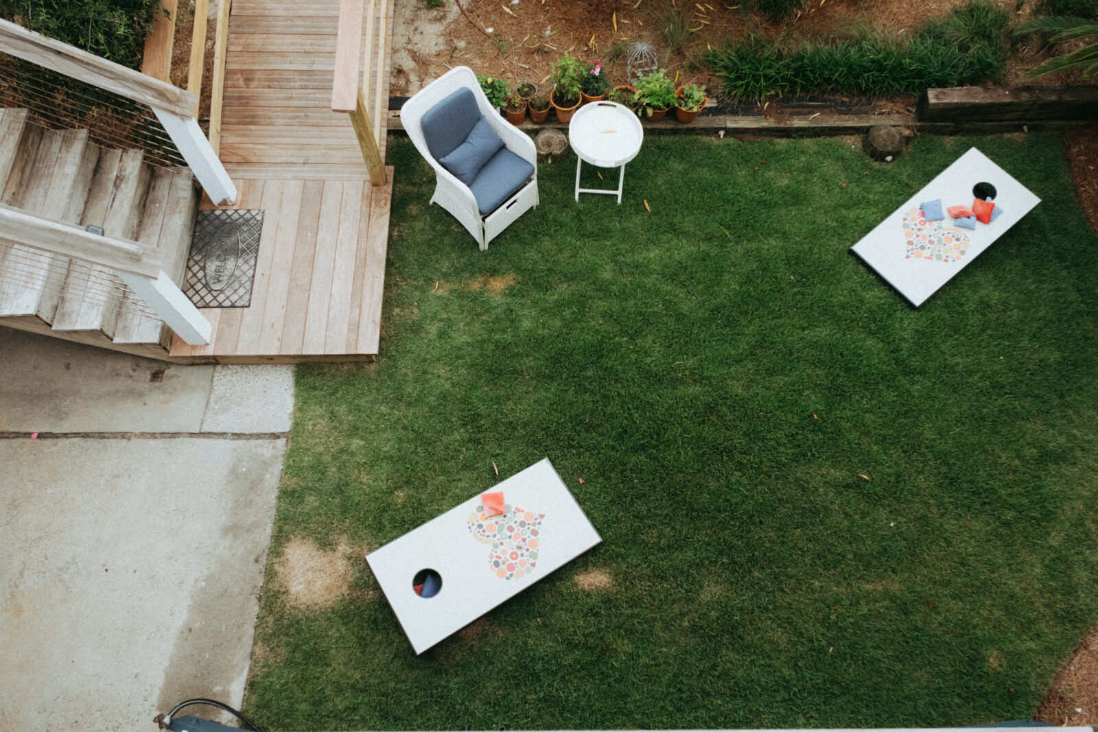 A backyard with green grass, two cornhole boards, a small white table, and a gray chair next to wooden steps leading down from a porch.