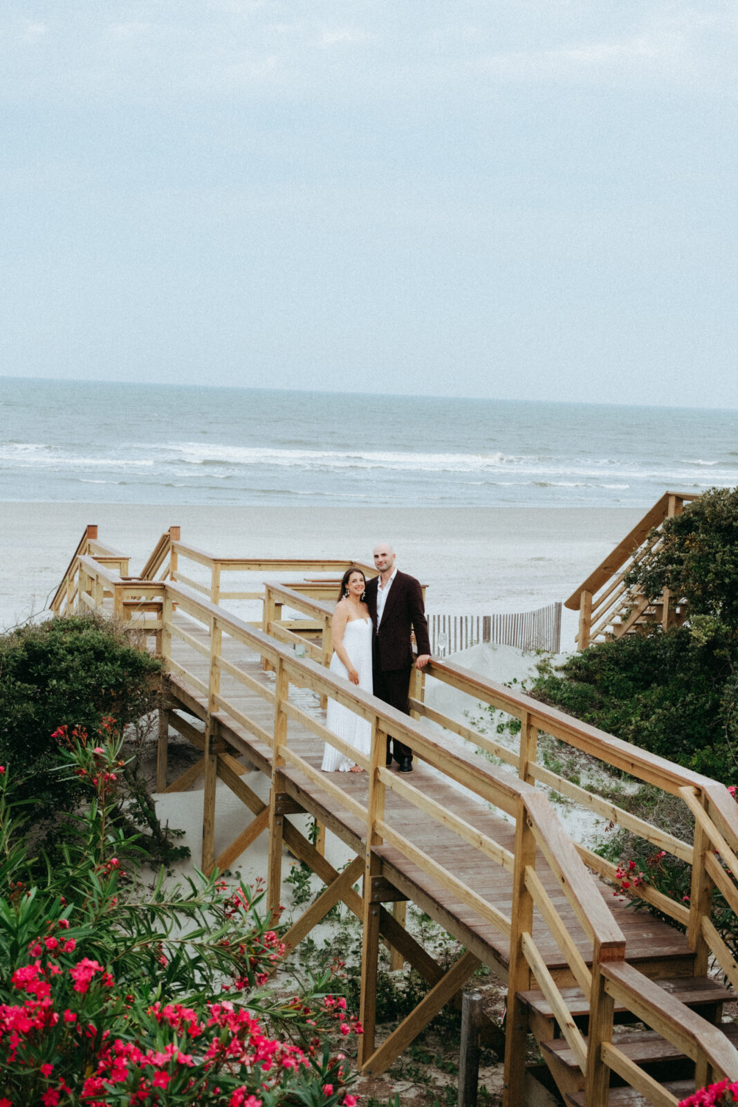A couple stands together on a wooden walkway leading to a sandy beach, with the ocean and waves in the background and greenery in the foreground.