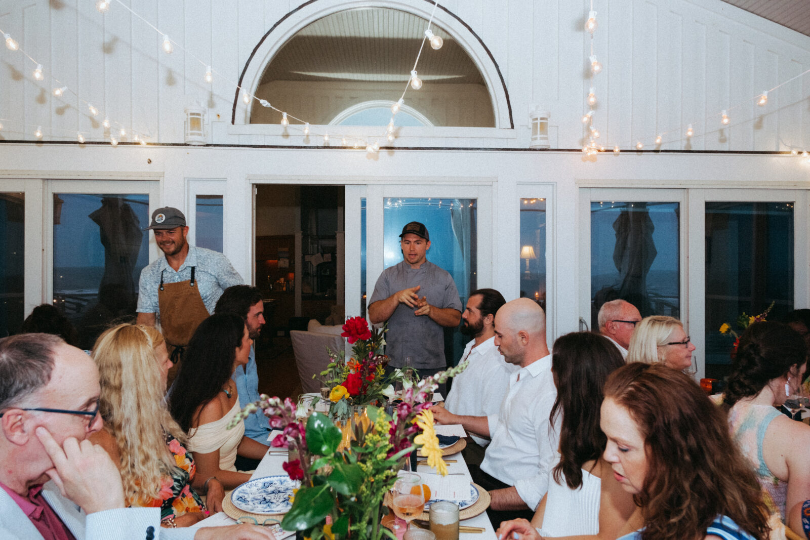 A group of people sit at a long, flower-adorned dinner table indoors while two men stand and address them; string lights hang above and windows are in the background.