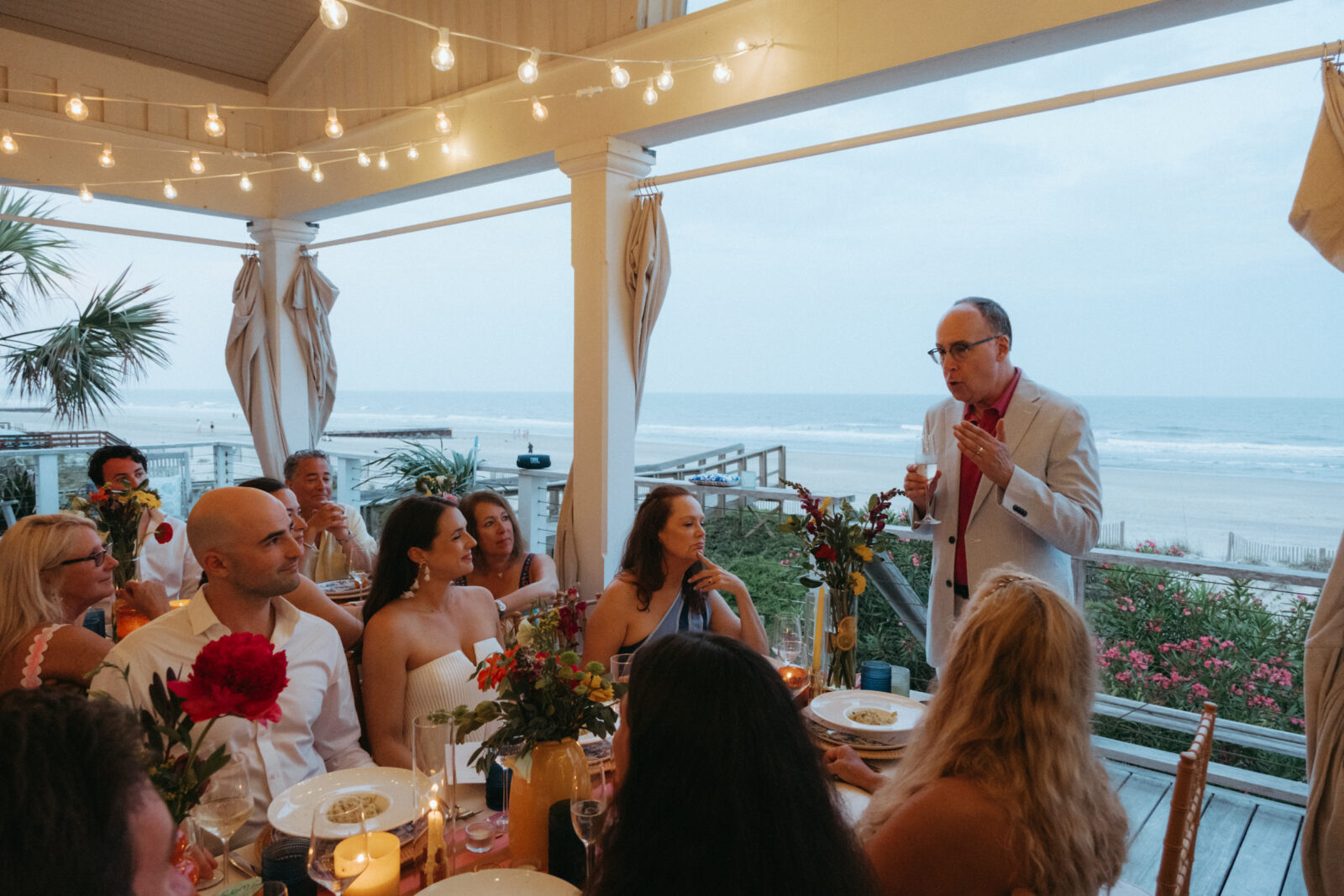 A man in a light suit stands and speaks to a seated group at an outdoor beachside dinner, with string lights overhead and the ocean visible in the background.
