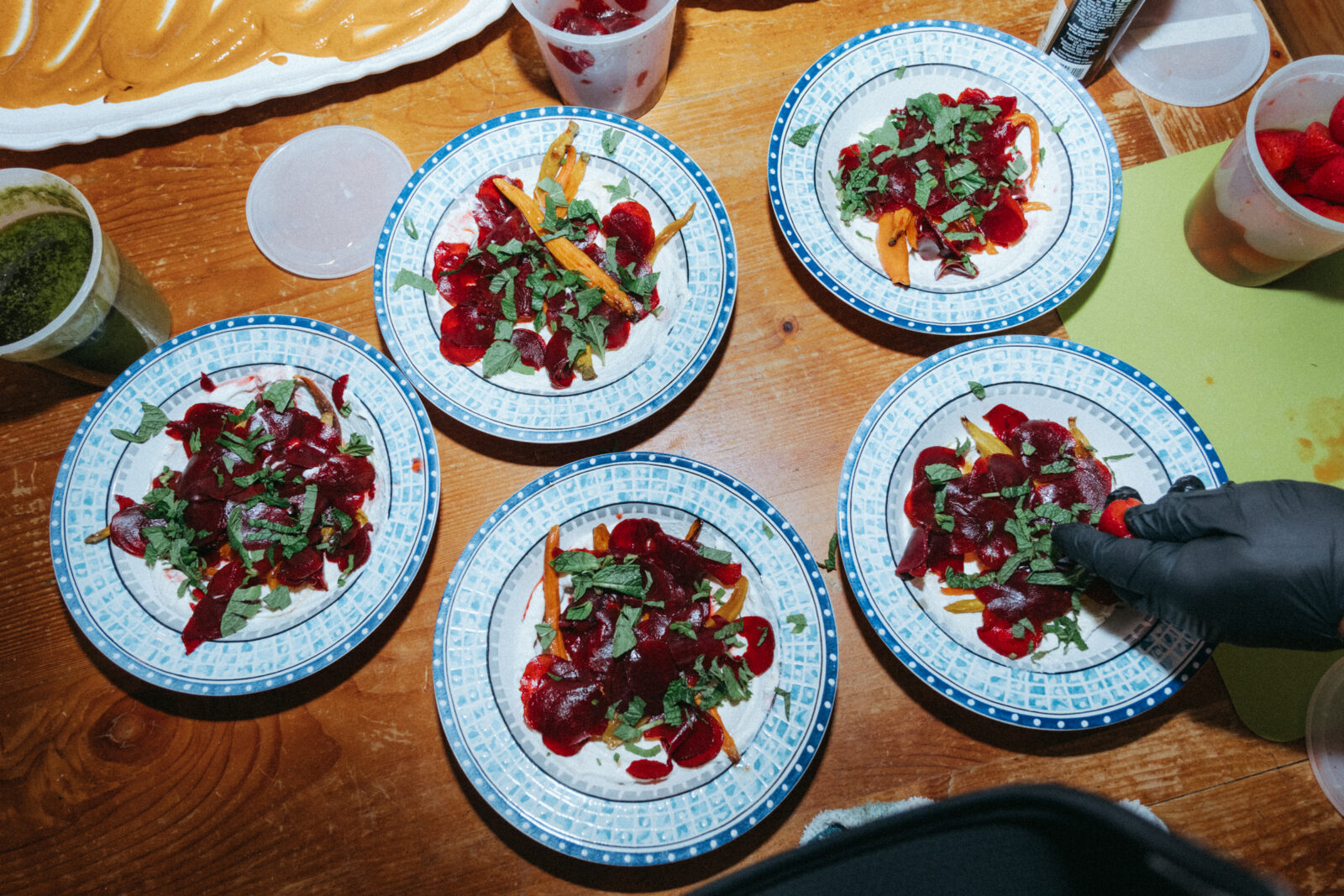 Five plates topped with sliced red and orange vegetables, greens, and herbs are arranged on a wooden table; a gloved hand garnishes one plate.