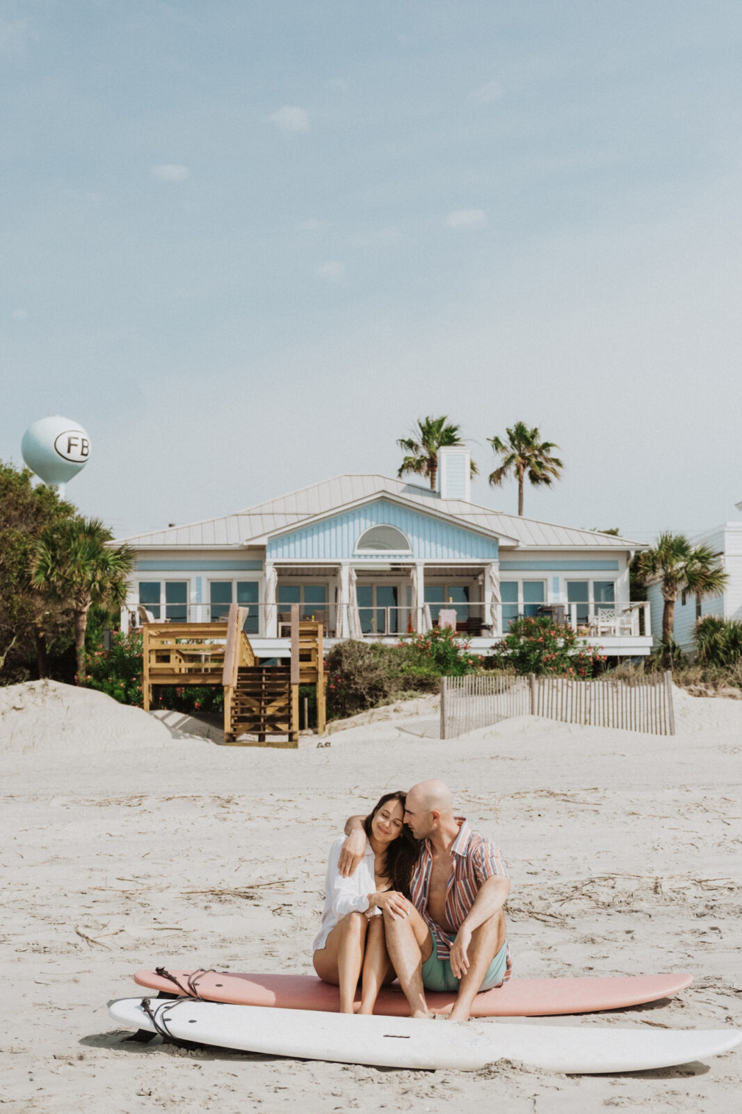 A couple sits on a surfboard on a sandy beach in front of a blue beach house, with palm trees and a balloon in the background.