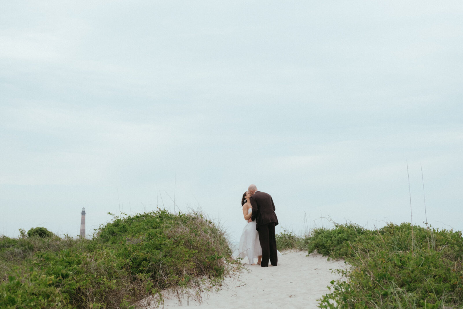 A couple in wedding attire stands on a sandy path between green bushes, embracing. A distant lighthouse is visible on the left under a cloudy sky.