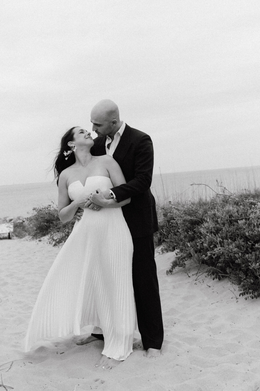 A couple dressed in formal attire stands on a sandy beach, embracing and smiling at each other, with the ocean and foliage visible in the background.