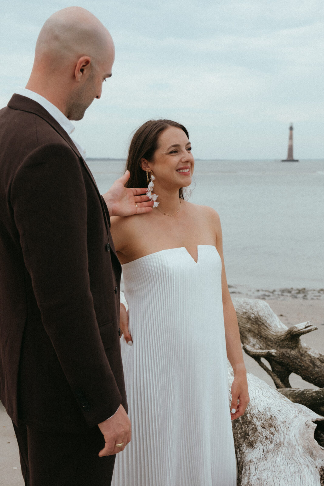 A couple stands on a beach; the man in a dark suit gently touches the smiling woman’s face. She wears a strapless white dress. A lighthouse is visible in the background over the water.