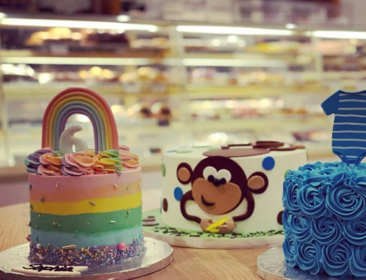 Three decorated cakes on a wooden table: a rainbow cake, a cake with a cartoon monkey face, and a blue cake with a shirt topper, with a bakery display case in the background.