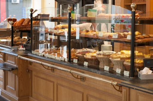Bakery counter displaying a variety of pastries, breads, and desserts behind a glass barrier with baskets and price tags visible.