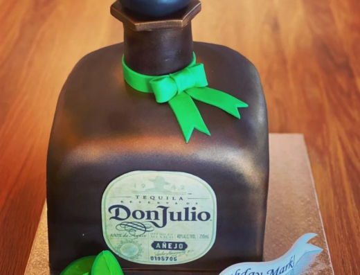 A cake shaped like a Don Julio tequila bottle with a green bow, lime slices, and a banner that reads, “Happy 40th birthday Mark!” on a wooden table.