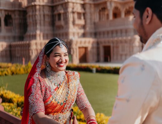 A woman in a red and gold saree holds hands with a man in a cream sherwani, standing outside an ornate temple on a sunny day.