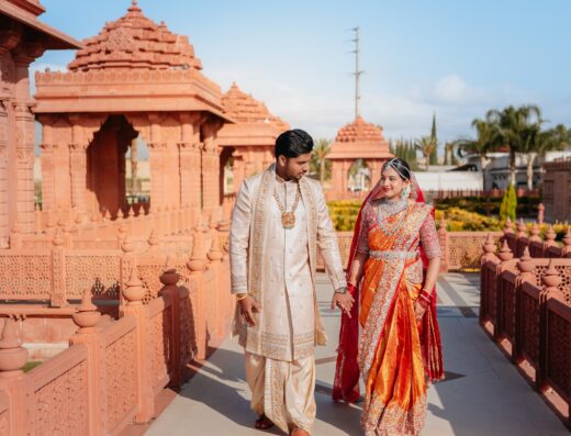 A couple in traditional Indian attire walks hand in hand at a temple with ornate red sandstone architecture under a clear blue sky.