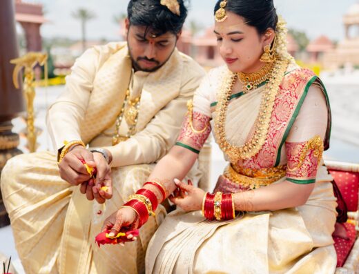 A couple in traditional Indian attire performs a wedding ritual with flower petals, seated outdoors beside bowls of flowers and ceremonial items.